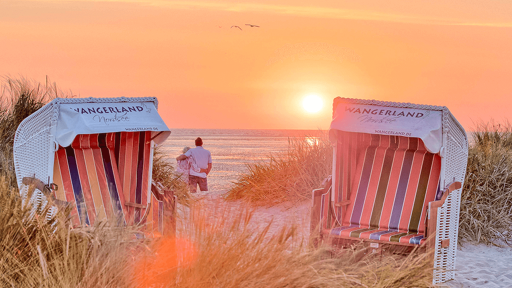 A Couple Of People Walking On A Beach With Chairs And A Sunset